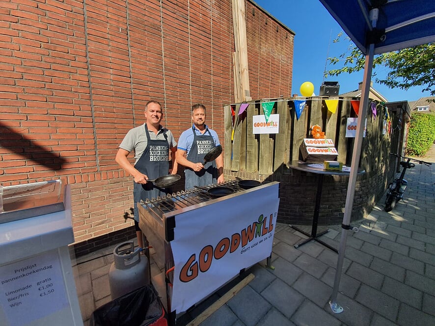 Two men in aprons standing behind a grill at an outdoor event, preparing food under a "GOODWILL" banner.