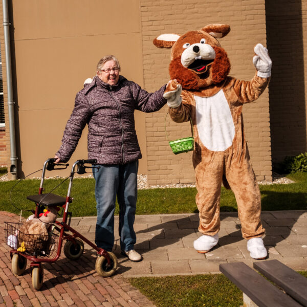 An elderly woman with a walker holding hands with a person in a brown dog mascot costume outside.