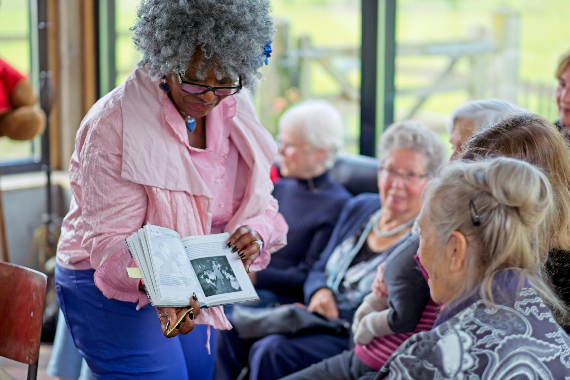 An elderly woman in pink showing a photo book to a group of older women seated in a bright room.