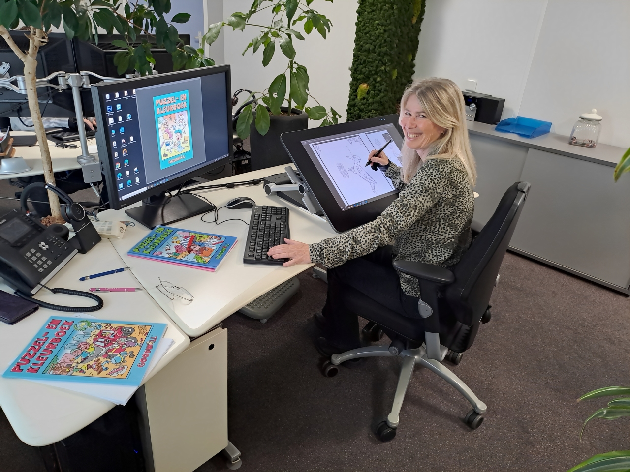 A smiling woman sitting at a desk, working on a digital drawing tablet with puzzle books on the table.